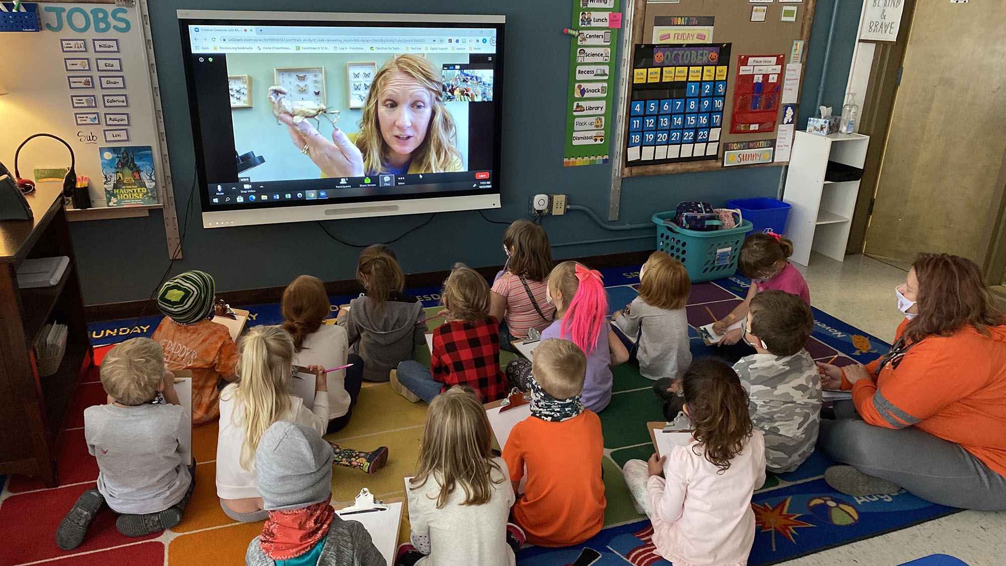 DistanceLearning Photo of MBHI Educator Carolyn holding a leaf insect on a screen while children sit on the floor and learn.